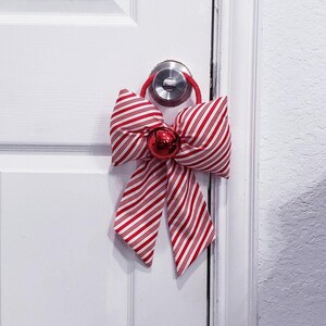 Peut inclure: Un accessoire de porte d&eacute;coratif compos&eacute; d'un grand n&oelig;ud en tissu &agrave; rayures rouges et blanches fa&ccedil;on sucre d'orge. Le n&oelig;ud est fix&eacute; &agrave; une boucle &eacute;lastique rouge qui s'adapte sur une poign&eacute;e de porte argent&eacute;e et comporte un grelot rouge brillant.