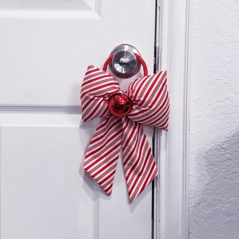 Peut inclure: Un accessoire de porte d&eacute;coratif compos&eacute; d'un grand n&oelig;ud en tissu &agrave; rayures rouges et blanches fa&ccedil;on sucre d'orge. Le n&oelig;ud est fix&eacute; &agrave; une boucle &eacute;lastique rouge qui s'adapte sur une poign&eacute;e de porte argent&eacute;e et comporte un grelot rouge brillant.