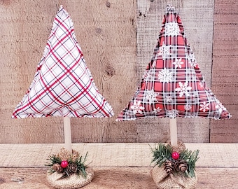 May include: Two miniature Christmas trees made of fabric with a plaid pattern. The trees are red and white with a burlap base and pine sprigs with red berries.