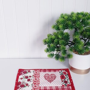 Puede incluir: Un posavasos de tela de patchwork rojo y blanco con un dise&ntilde;o de coraz&oacute;n en el centro. El posavasos est&aacute; rodeado por un borde rojo con peque&ntilde;os corazones rosas.