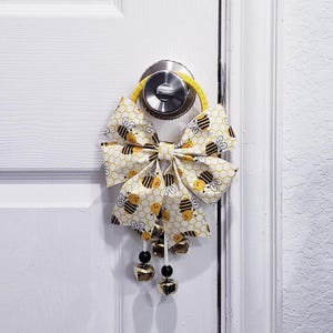 以下が含まれることがあります： ハチをテーマにした装飾用のドアリボン。リボンは、ハニカム模様と漫画のハチが描かれた、黄色と白の生地で作られています。ドアノブに掛けられ、金色のベルと黒いビーズが付いています。装飾品です。