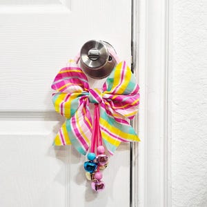 May include: A pink, yellow, and blue striped bow with a pink ribbon and jingle bells hanging from a white door.
