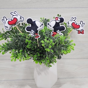 Könnte beinhalten: Eine weiße Keramikvase mit grünen, belaubten Pflanzen. Drei Stofffiguren von Mickey Mouse und Minnie Mouse sind an Holzspießen befestigt und in der Vase platziert. Die Figuren küssen sich und halten Herzen.