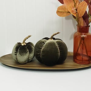 May include: Two olive green velvet pumpkins with metallic gold stems, positioned on an oval wooden tray next to a clear amber glass vase containing orange and brown faux foliage. The pumpkins feature a soft, plush texture and ribbed detailing.