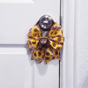 May include: A decorative door hanger featuring a fabric bow with a yellow and brown sunflower pattern. The bow is centered with a large, dark metallic jingle bell and is attached to a circular loop designed to fit over a standard round door knob.