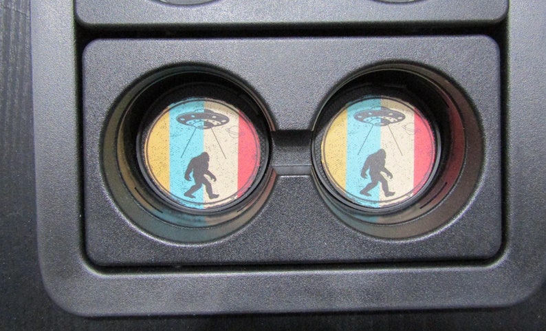 May include: Two round drink coasters with a retro design featuring a Bigfoot walking under a flying saucer. The coasters are yellow, blue, and red with a white background.