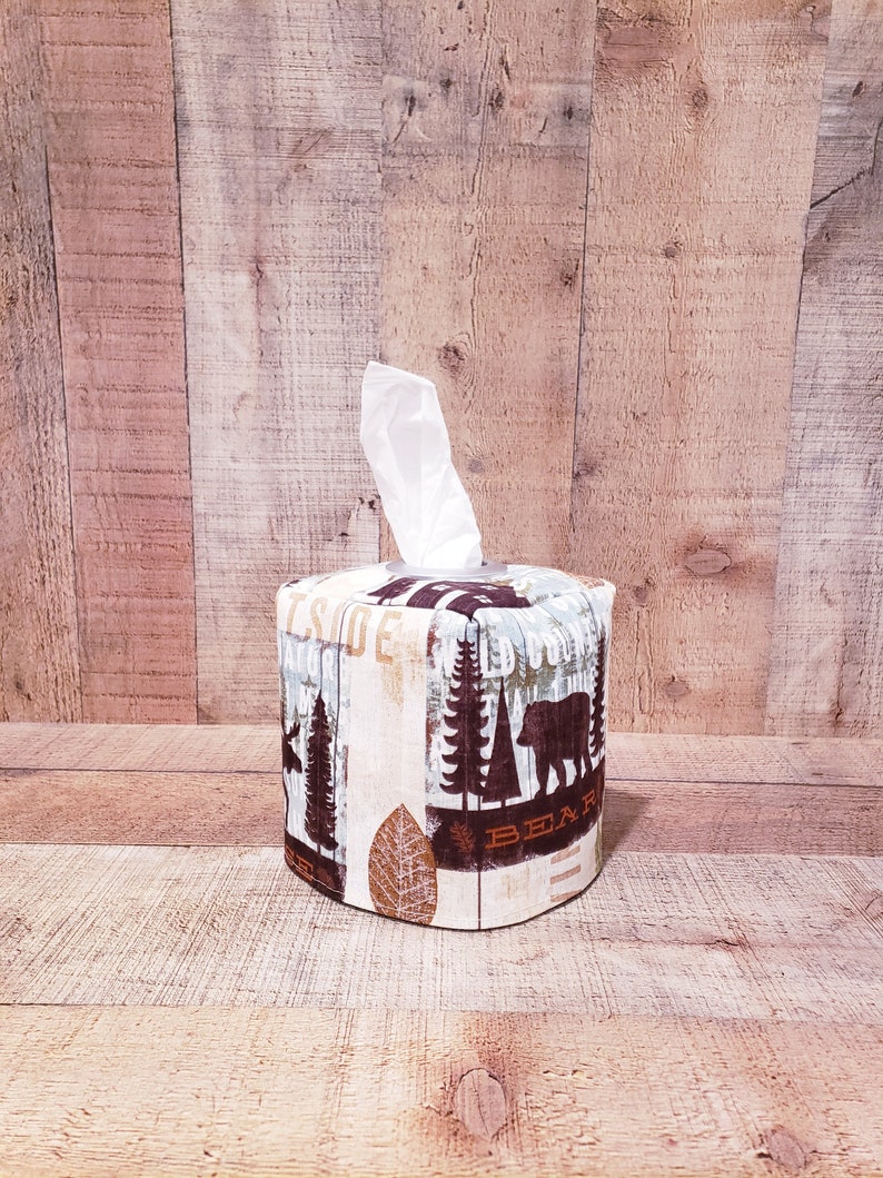 Op de afbeelding: Een bruine en witte tissueboxhoes met een bosontwerp met bomen, een beer en het woord "Beer".