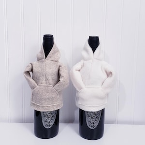 May include: Two wine bottle covers shaped like hoodies. One is a light beige color, and the other is white. Each has a hood and a front pocket. The bottles are black with a decorative label.