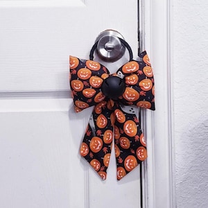 Könnte beinhalten: Eine Stoffschleife mit einem Muster aus orangefarbenen Kürbissen auf schwarzem Hintergrund. Die Schleife ist an einem schwarzen Gummiband befestigt und hat in der Mitte eine große, runde, mattschwarze Glocke als zentrales Element.