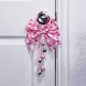 以下が含まれることがあります： ハート模様のピンクと白のリボンがドアノブから吊り下げられています。弓には、ぶら下がっているピンクとシルバーのベルが飾られています。ドアは白です。