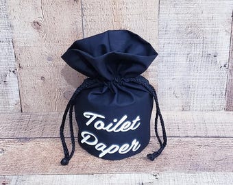 May include: A navy blue drawstring bag with the words "Toilet Paper" in white script. The bag is gathered at the top and has a black drawstring closure. The background is a light brown wood.