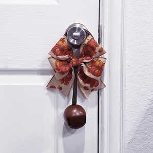 May include: A decorative door hanger featuring a large bow made of tan fabric with an orange and brown pumpkin print. A dark brown ribbon hangs from the center of the bow, attached to a large, round, dark brown metal jingle bell at the bottom.
