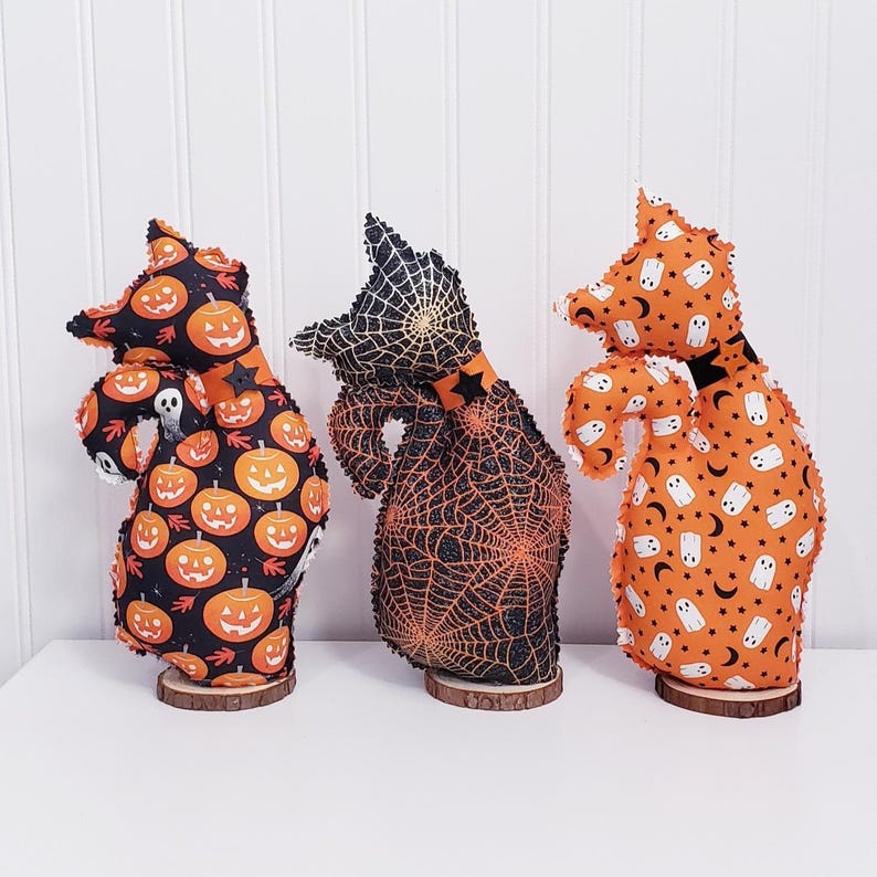 May include: Three Halloween-themed fabric figures on wooden bases. The figures have different patterns: pumpkins on black, a spiderweb, and ghosts with moons and stars on orange. Each figure has a pointed hat and a curved body.