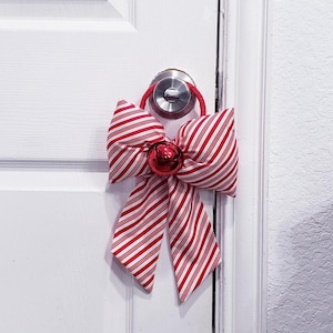 Könnte beinhalten: Eine dekorative Stoffschleife mit rot-weißem Zuckerstangen-Streifenmuster. Die Schleife ist an einer roten Schlaufe befestigt und hat eine einzelne, glänzende rote Glocke in der Mitte, die zum Aufhängen an einem runden Türgriff gedacht ist.
