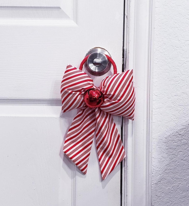 Peut inclure: Un n&oelig;ud d&eacute;coratif en tissu avec un motif ray&eacute; rouge et blanc rappelant les sucres d'orge. Le n&oelig;ud est fix&eacute; &agrave; une boucle rouge et comporte une clochette rouge brillante au centre, con&ccedil;u pour &ecirc;tre suspendu &agrave; une poign&eacute;e de porte ronde.