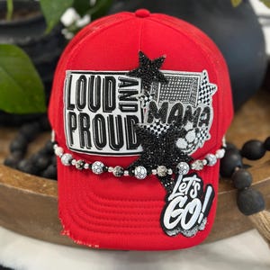 May include: A bright red trucker hat featuring a white patch with the black text LOUD AND PROUD, a black rhinestone star, a soccer-themed patch with the word MAMA, and a white patch with the black text LET'S GO! A beaded trim with soccer ball accents.