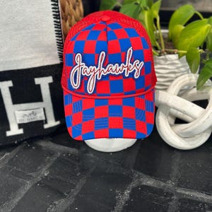 May include: A red and blue checkered baseball cap with the word "Jayhawks" in script across the front. The cap has a mesh back and a curved brim. The hat is displayed on a white stand.