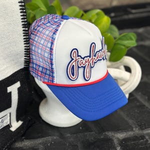 May include: A white and blue trucker hat with a blue brim and a red and blue plaid pattern on the top. The word "Jayhawks" is embroidered on the front in a script font with red and blue accents.