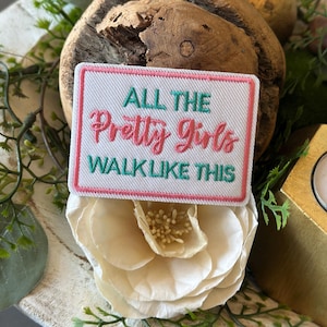 May include: A white patch with a pink border and the text "ALL THE Pretty Girls WALK LIKE THIS" in green and pink lettering. The patch is placed on a white flower and a wooden decorative piece.