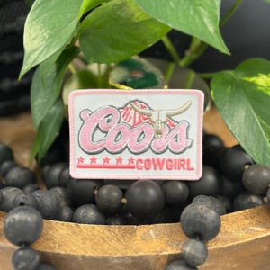 May include: A rectangular embroidered patch with a light pink border. The patch features the word Coors in pink script with a white outline, a white steer skull graphic, and the word COWGIRL in pink capital letters above five pink stars.