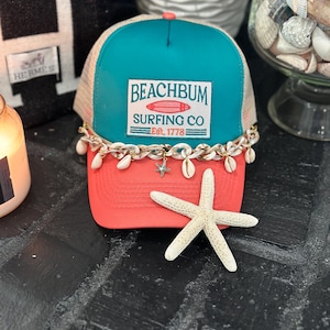 May include: A teal and coral trucker hat with the text "Beachbum Surfing Co Est. 1778". The hat features a mesh back, a chain with seashells, and a starfish. The hat is a beach-themed accessory.