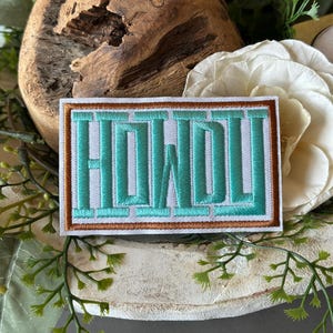 May include: Embroidered patch with the word "HOWDY" in turquoise block letters. The patch has a white background with a brown border. The patch is rectangular and is surrounded by greenery, a wooden decorative piece, and a white flower.