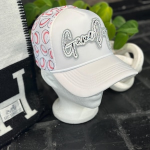May include: White baseball cap with a mesh panel featuring a pattern of red baseballs. The cap has the text "Game Over" in silver and rhinestone embellishments. The cap is displayed on a white mannequin head.