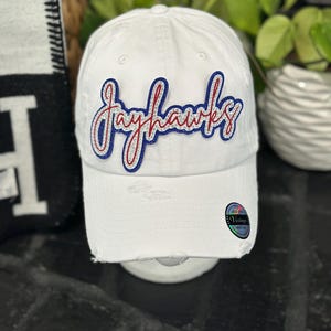 May include: White baseball cap with distressed details. The cap features the word "Jayhawks" in a script font with red and blue accents. A small, circular tag is visible on the side of the cap.