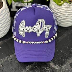 May include: Purple trucker hat with a white and purple checkered pattern on the top and a curved brim. The hat features the text "Game Day" in silver and white, with "WILDCATS" spelled out on the brim.