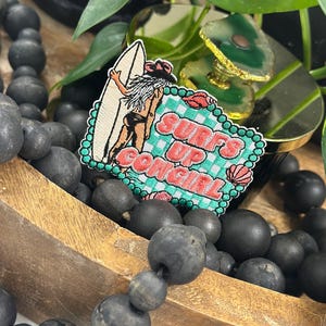 May include: A rectangular embroidered patch featuring a person with long hair wearing a hat and holding a surfboard. The design includes a green and white checkered background with pink seashells and the text SURFS UP COWGIRL in pink letters.