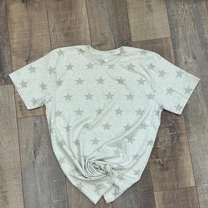May include: A short-sleeved crew neck t-shirt in a light heathered beige color. The garment features an all-over repeating pattern of five-pointed stars in a slightly darker shade of beige, distributed evenly across the front and sleeves.
