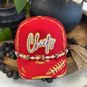May include: A red trucker hat featuring the word Chiefs in white and gold script lettering. The hat is decorated with a beaded strand of white pearls and miniature footballs across the front, with gold glitter accents on the brim and crown.