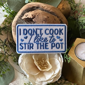 May include: Embroidered patch with the text "I DON'T COOK I like to STIR THE POT" in blue lettering on a white background. The patch is rectangular with a blue border and small heart accents. The patch is displayed on a wooden bowl with floral and greenery accents.