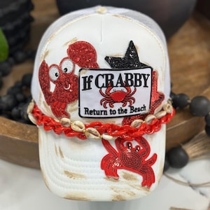 May include: A white trucker hat featuring red sequined crab appliqués and a rectangular patch with the text If CRABBY Return to the Beach. The hat is accented with a red plastic chain and small tan seashells across the front panel.