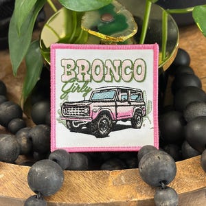 May include: A square embroidered patch with a pink border on a white background. The patch features a pink SUV illustration, green cactus accents, and the text "BRONCO Girly" in a stylized green and pink font. The design is centered and clear.