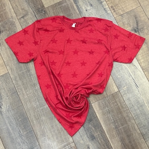 May include: A short-sleeved crew neck t-shirt in a vibrant red color. The fabric features a repeating pattern of tonal, slightly darker red stars printed across the entire garment, including the sleeves and the main body of the shirt.