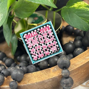 May include: A square embroidered patch featuring a black and white checkered background with the pink text "SMALL TOWN SMOKE SHOW" in the center. The patch is bordered by a bright turquoise stitched edge with a repeating dot pattern.