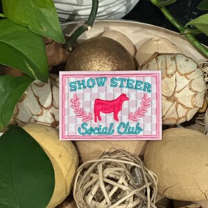 May include: A rectangular patch with the text "SHOW STEER Social Club" in teal and white, and a pink steer illustration. The patch has a pink and white gingham border and is surrounded by decorative elements.