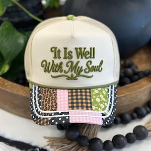 May include: A cream-colored trucker hat featuring the embroidered green text 'It Is Well With My Soul' on the front panel. The brim displays a patchwork pattern of leopard print, pink gingham, black and tan plaid, and green floral fabric.