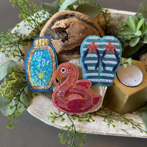 May include: Embroidered beach-themed patches featuring a blue sunscreen bottle with "SPF 50+" text, a pink flamingo, and a pair of blue and white striped flip-flops with red starfish accents. The patches are arranged in a decorative wooden bowl with greenery.
