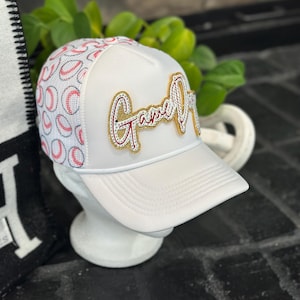 May include: White baseball cap with "Game On" text in gold and red embellishments. The cap features a white mesh back with a baseball pattern. A stylish accessory for sports enthusiasts.