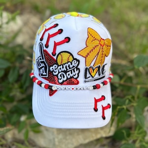 May include: White baseball cap with a mesh back, featuring red and yellow embroidered accents. The cap has the words "Game Day" and "Play Ball" on it, along with a yellow bow and a softball design.