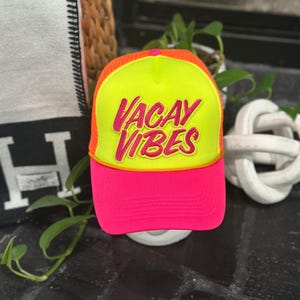 May include: A neon pink and orange trucker hat with the words "VACAY VIBES" in pink script. The hat has a yellow front panel and a pink brim. The text is outlined in a gold rope-like trim.
