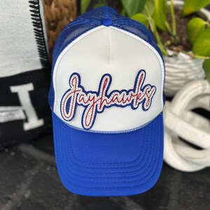 May include: A blue and white trucker hat with the word "Jayhawks" in red script. The hat has a blue brim and a white front panel with a blue mesh back. The text is outlined in red and blue.
