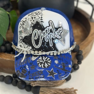 May include: A blue and white trucker hat featuring a black sequined star with the text Cheer Mom in white script. The front panel includes a leopard print section, a rhinestone trim, and a white bow. The brim displays blue and black cheer graphics.