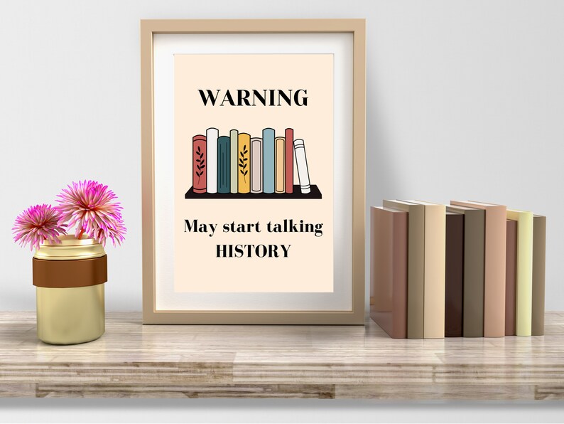 May include: A framed print with the text "WARNING May start talking HISTORY" above a shelf of illustrated books. A vase with pink flowers sits to the left, and a stack of books is on the right. The frame is a light brown colour.
