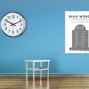 May include: A round wall clock with a silver rim and black hands is mounted on a blue wall. To the right is a white poster with the text "DEAD WORDS" and a tombstone graphic listing overused words.