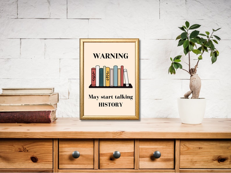 May include: A gold framed print with a beige background. The print features a warning sign with the text "WARNING" and a row of colorful books with the text "May start talking HISTORY".