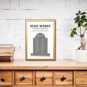 May include: A framed print with the words "DEAD WORDS" in black text. The print features a gray tombstone with a list of overused words. The frame is gold, and the print is displayed on a wooden surface with a plant and books.