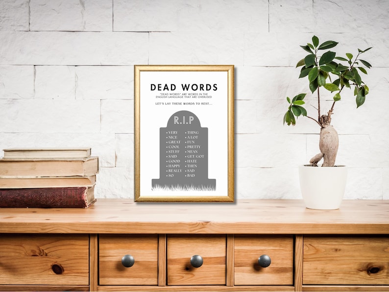 May include: A framed print with the words "DEAD WORDS" in black text. The print features a gray tombstone with a list of overused words. The frame is gold, and the print is displayed on a wooden surface with a plant and books.